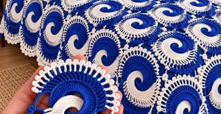 A crocheted bedspread made from blue and white spiral motifs with scalloped edges, draped over a bed, with a hand holding a single crochet motif in the foreground.