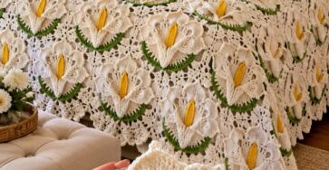 A crocheted bedspread with white calla lily flower motifs, green leaves, and yellow centers on a bed. Hands hold one of the floral crochet motifs in the foreground.
