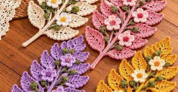 Four crochet fern leaf mats in cream, pink, purple, and yellow, each adorned with green buds and 3D flowers, displayed on a wooden tabletop.
