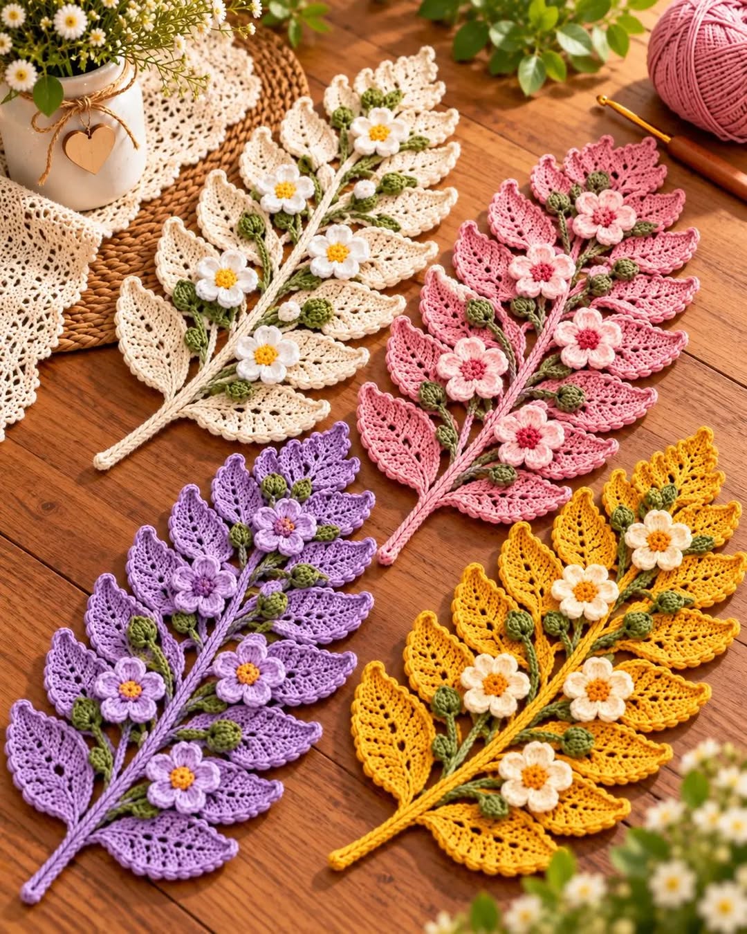 Four crochet fern leaf mats in cream, pink, purple, and yellow, each adorned with green buds and 3D flowers, displayed on a wooden tabletop.