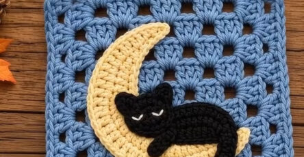 A crochet granny square featuring a blue background with a yellow crescent moon and a sleeping black cat appliqué.