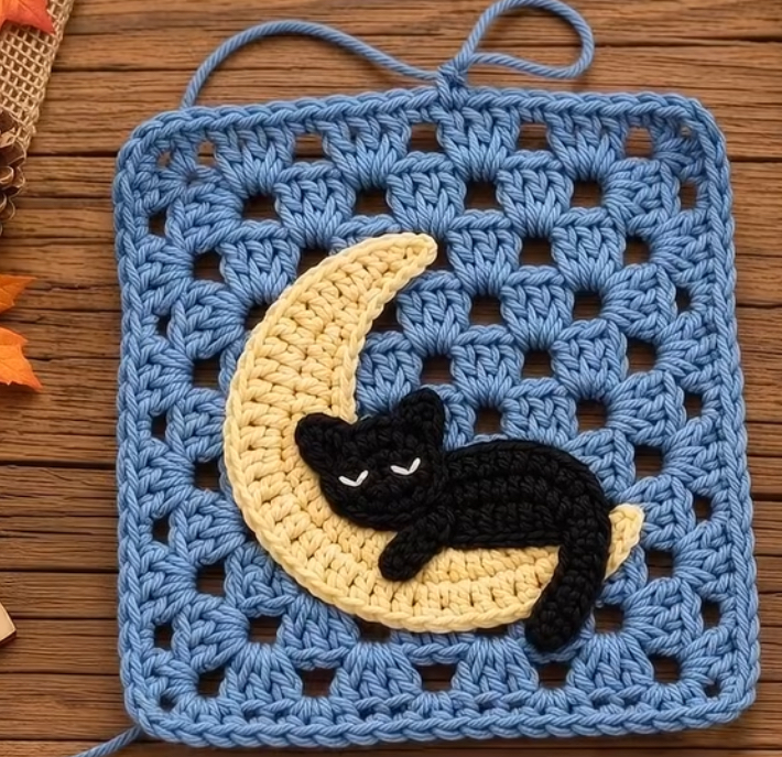 A crochet granny square featuring a blue background with a yellow crescent moon and a sleeping black cat appliqué.