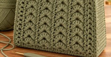 A sage green crochet handbag with textured panels, sturdy handles, and a fabric-lined interior, displayed with a ball of yarn and a crochet hook.