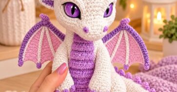 A crocheted amigurumi baby dragon in cream and purple with lavender accents and large, expressive eyes.