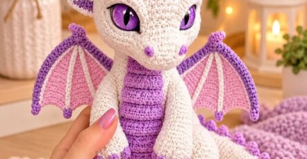 A crocheted amigurumi baby dragon in cream and purple with lavender accents and large, expressive eyes.